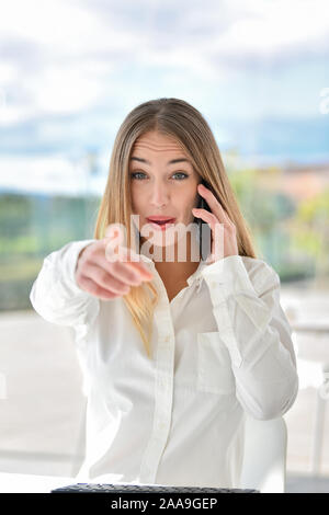 Smiling young woman having a conversation using smart phone over with finger to the camera and to you, hand sign, positive and confident gesture. Faci Stock Photo