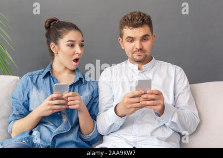 Relationnship Concept. Young couple woman with smartphone looking shocked at man browsing media sitting on sofa studio isolated on grey Stock Photo