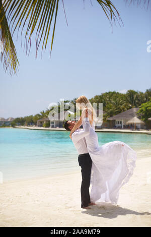 Happy groom holds bride on his hands under wedding ceremony arch on tropical island beach on Maldives. Turquoise ocean lagoon, white sand and Stock Photo