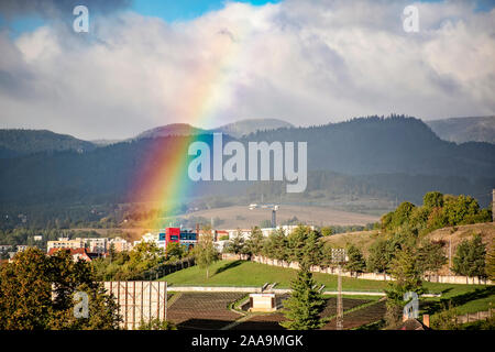 End of a rainbow in the sky over block of flats in the city with mountains behind. Cityscape Banska Bystrica, Slovakia Stock Photo