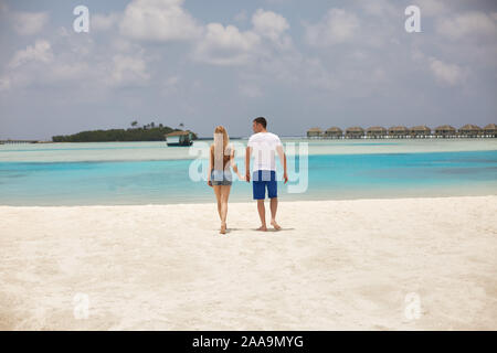 Back view of couple holding hands and walking to the blue ocean lagoon on Maldives at luxury spa resort. Travel and honeymoon concept. Stock Photo