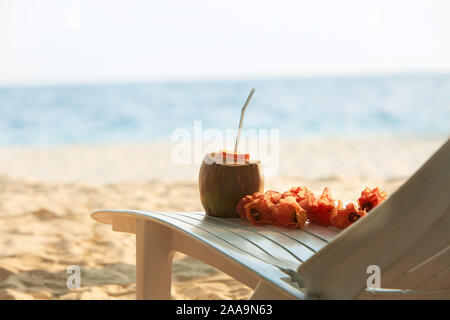 Coconut with drinking strawand lei flowers on beach bench or deck chair with blue ocean and white sand on background. Maldives Stock Photo