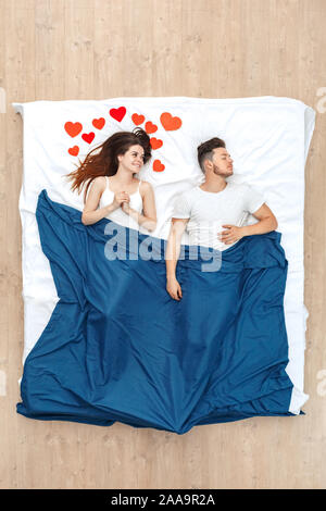 Bedtime. Young woman looking at sleeping man in love surrounded by hearts lying under blanket on bed top view Stock Photo