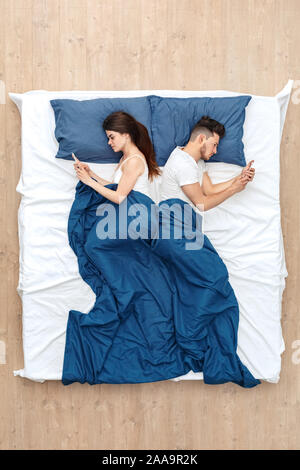 Bedtime. Young couple lying under blanket on bed back to back top view browsing smartphone antisocial Stock Photo
