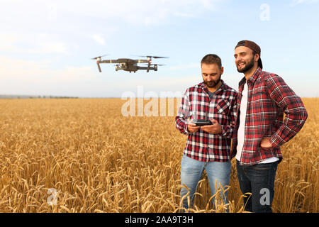 Compact drone hovers in front of two hipster men. Quadcopter flies near farmer and agronomist exploring harvest with innovative technology taking Stock Photo