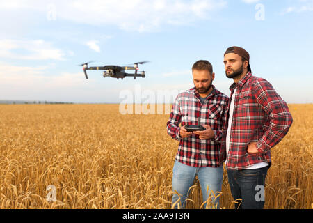Compact drone hovers in front of two hipster men. Quadcopter flies near farmer and agronomist exploring harvest with innovative technology taking Stock Photo