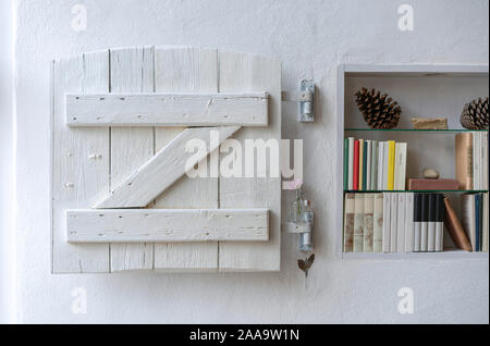 Detail of a small decorated bookshelf with white wooden shutter in a white wall Stock Photo