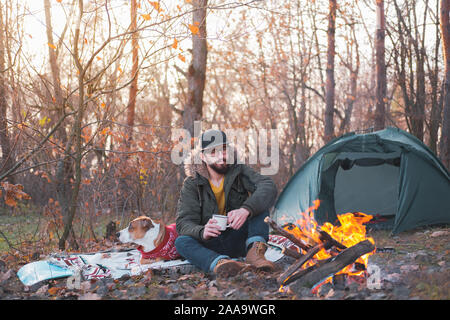 Bearded man enjoys company of his dog outdoors at a camping place in late autumn. Spending leisure time alone with a pet: thoughtful male person sits Stock Photo