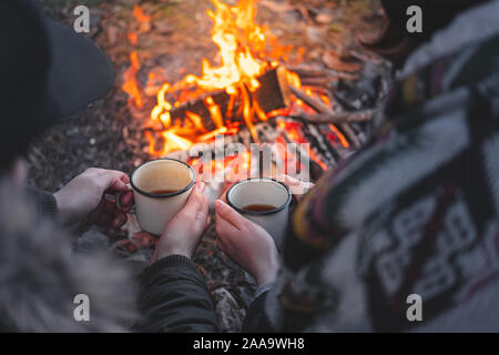 Two people warming hands with hot drinks by the bonfire. Spending nice time outdoors in chilly weather at a camping place - tranquil and peaceful scen Stock Photo
