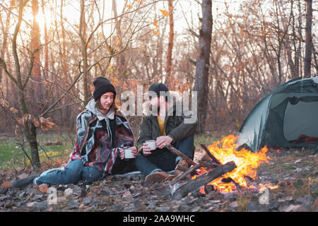Two persons enjoying hot drinks by the bonfire at a camping site. Going on a trekking vacation at cold season: man and woman sit by the fire with a te Stock Photo