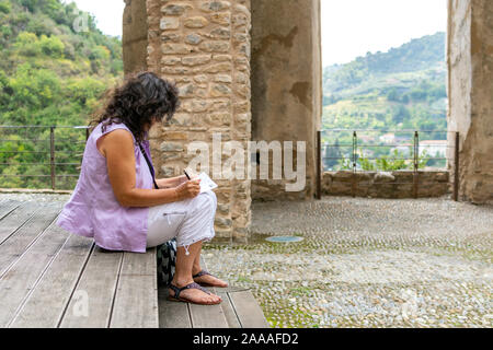 A female traveler writes postcards as she sits inside the ancient castle in the village of Dolceacqua, Italy. Stock Photo