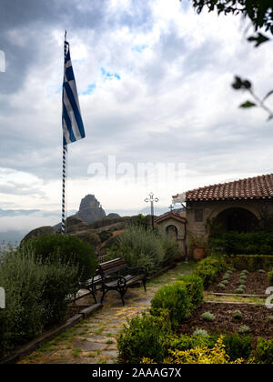 Stone building of the Holy Monastery of St. Stephen with terracotta tile roof, gardens, Greek flag, and cloudy valley with rugged mountain in the back Stock Photo