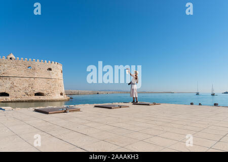 Happy tourist girl on holiday trip to Heraklion, Crete, Greece. Young woman traveller enjoyes sunshine sitting on the dock of Heraklion Venetian port Stock Photo