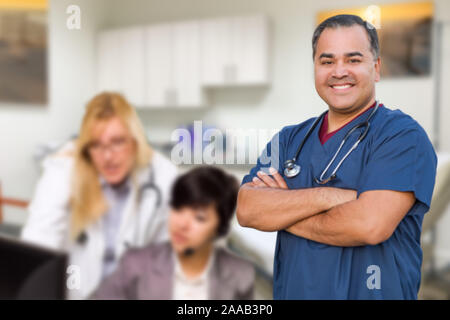 Handsome Hispanic Doctor or Nurse Standing in His Office with Staff Working Behind. Stock Photo