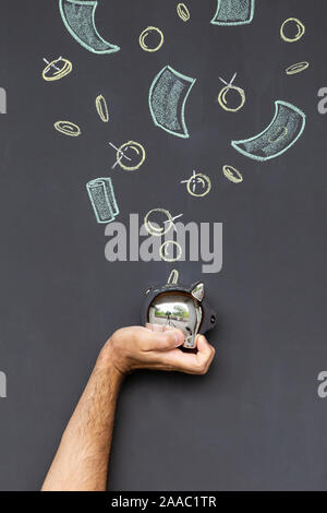 Concept of saving money with a silver piggy bank held in a hand in front of a blackboard with hand drawn coins and banknotes Stock Photo