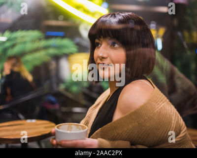 Beautiful young girl sits and drinks coffee at a trendy cafe or bar at night with window reflection.View through window glass. Stock Photo