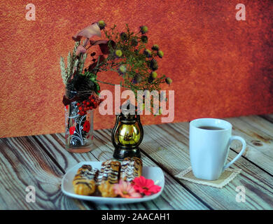Warm still life, a plate with eclairs, a mug of tea on a napkin, an autumn herbarium and an old-fashioned lamp. Close-up. Stock Photo