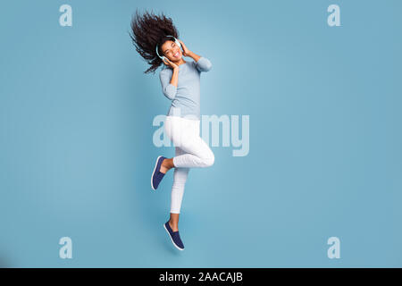 Turned full length body size photo of cheerful positive cute nice sweet girl listening to music in headphones jumping with hair flying isolated pastel Stock Photo