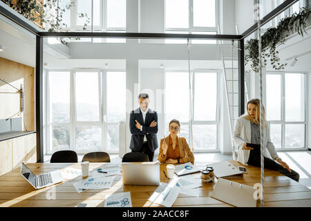 Group of a young office employees having some office work at the large meeting table, wide view on the spacious room with large windows Stock Photo