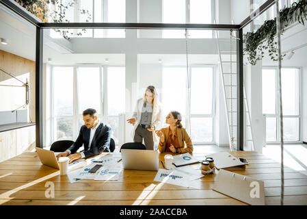 Group of a young office employees having some office work at the large meeting table, wide view on the spacious room with large windows Stock Photo