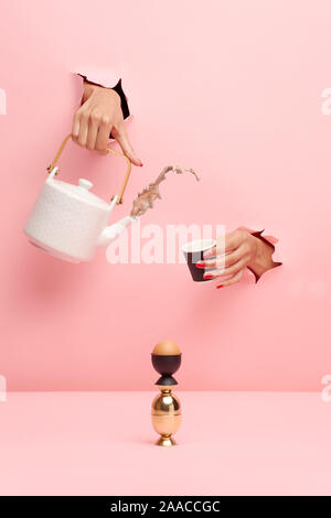 Women's hand braking through the pink background and holding a tea pot and a cup. An egg is set on the table. Stock Photo
