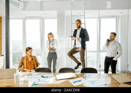 Group of a young creative office employees having some informal discussion, while standing together in the bright meeting room with large windows Stock Photo
