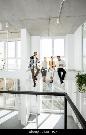 Group of a young creative office employees having some informal discussion, while standing in the two-level bright meeting room with large windows Stock Photo