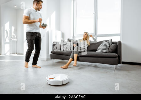 Young couple relaxing on the couch while automatic vacuum cleaner doing the housework in the modern white living room. Household robots concept Stock Photo