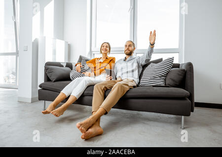 Happy young couple sitting together on the couch in the bright living room of the modern apartment, enjoying their new home Stock Photo