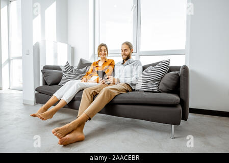 Happy young couple using digital tablet while sitting together on the couch in the bright living room of the modern apartment Stock Photo