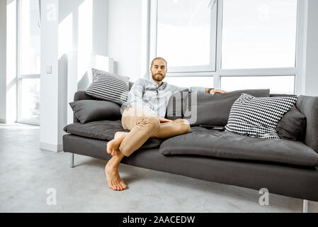 Full length portrait of a handsome caucasian man sitting on the couch in the bright living room at the modern apartment Stock Photo
