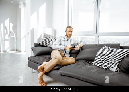 Full length portrait of a handsome caucasian man sitting with digital tablet on the couch in the bright living room Stock Photo
