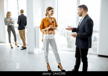 Young elegantly dressed people meeting in the white hallway or showroom, talking and drinking wine during some informal event Stock Photo