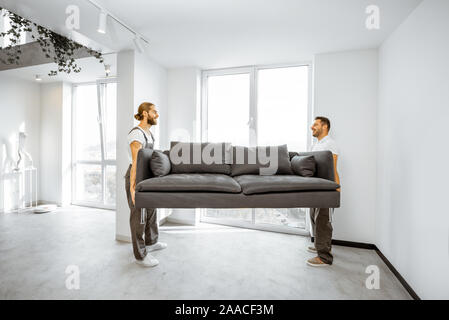 Two professional movers in workwear placing large couch in the living room of the modern white apartment. Furniture delivery concept Stock Photo