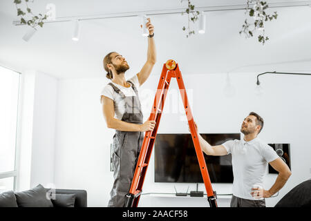 Two repairmen or professional electricians installing light spots, standing on the ladder in the living room of the modern apartment Stock Photo