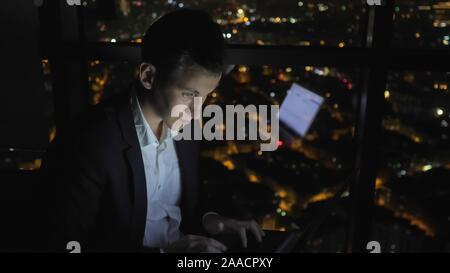 Young man freelancer is working on his computer near the window in home at night. Self-employed man has overtime job. Freelance and self-employed concept. Stock Photo