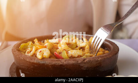 Good served food in restaurant. Woman is eating frying hot shrimps with vegetables served in clay bowl, closeup dish. Takes shrimp out of the boiling oil with a fork. Stock Photo