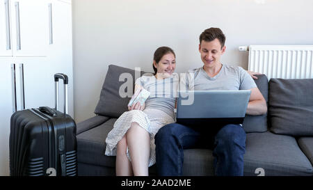 Young couple is booking transfer to the airport on laptop sitting on sofa at home. They are going on vacation with passports and suitcase. Stock Photo