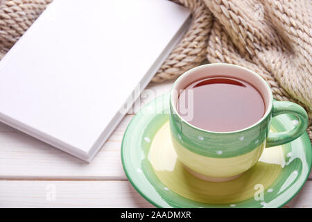 Still life with cup of tea or coffee and white book next to winter woolen scarf. White wooden table top. Space for your text. Stock Photo