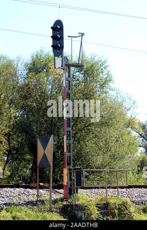 Tall railway tracks traffic light mounted on strong metal pole next to old dilapidated dirty sign and safety fence surrounded with uncut grass Stock Photo