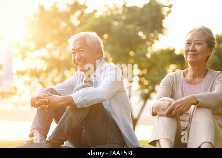 asian senior couple sitting on grass enjoying sunset outdoors in park Stock Photo