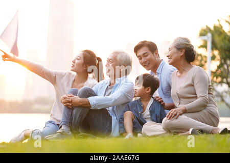 three generation happy asian family sitting on grass enjoying good time at dusk outdoors in park Stock Photo
