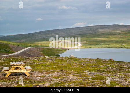 Wooden picnic table with benches near a lake and empty asphalt road through summer norwegian green hills in Finnmark, Norway. Stock Photo