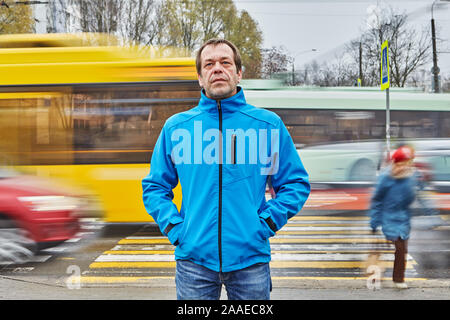 A sad middle-aged man, over 50 years old, dressed in a blue jacket, stands in front of a pedestrian crossing with his hands in pockets, probably he is Stock Photo