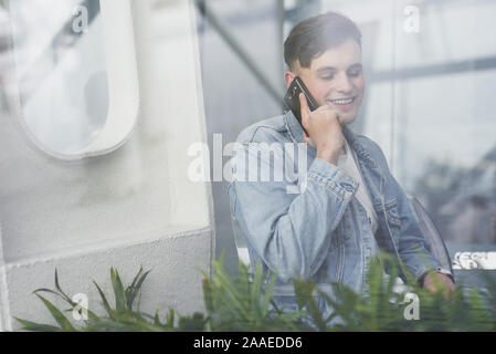 A smiling young man speaks by telephone in a cafe, a view from the street Stock Photo