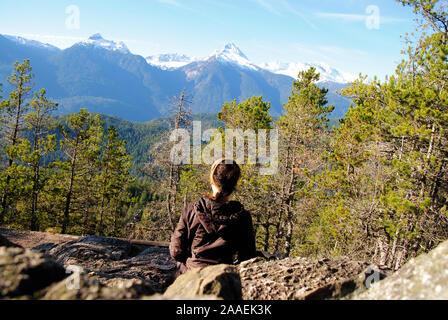 A natural young woman with brown braided hair, sitting still on rocks in a contemplative posture, staring at the snow capped Tantalus mountain range Stock Photo