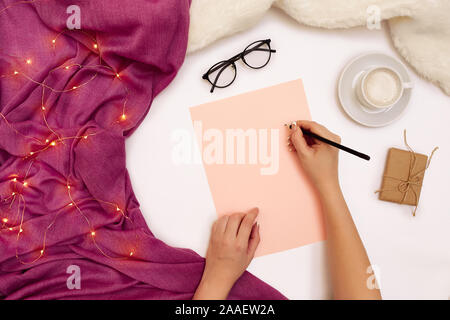 Top view of girl's hand writing in notepad placed on white surface with coffee cup and other items. Mock up Stock Photo