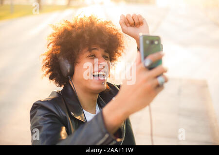 Handsome young black man with afro hairstyle listening music with mobile and headphones in a sunny city Stock Photo