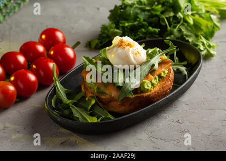 bruschetta salmon with avocado - salted salmon with guacamole, arugula and poached egg on a warm fried bun in a black plate on a gray background Stock Photo