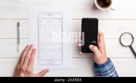 Designer tests the user interface and user experience on a mobile phone. Wireframe beside with mobile phone with a sketched app. The concept of design Stock Photo